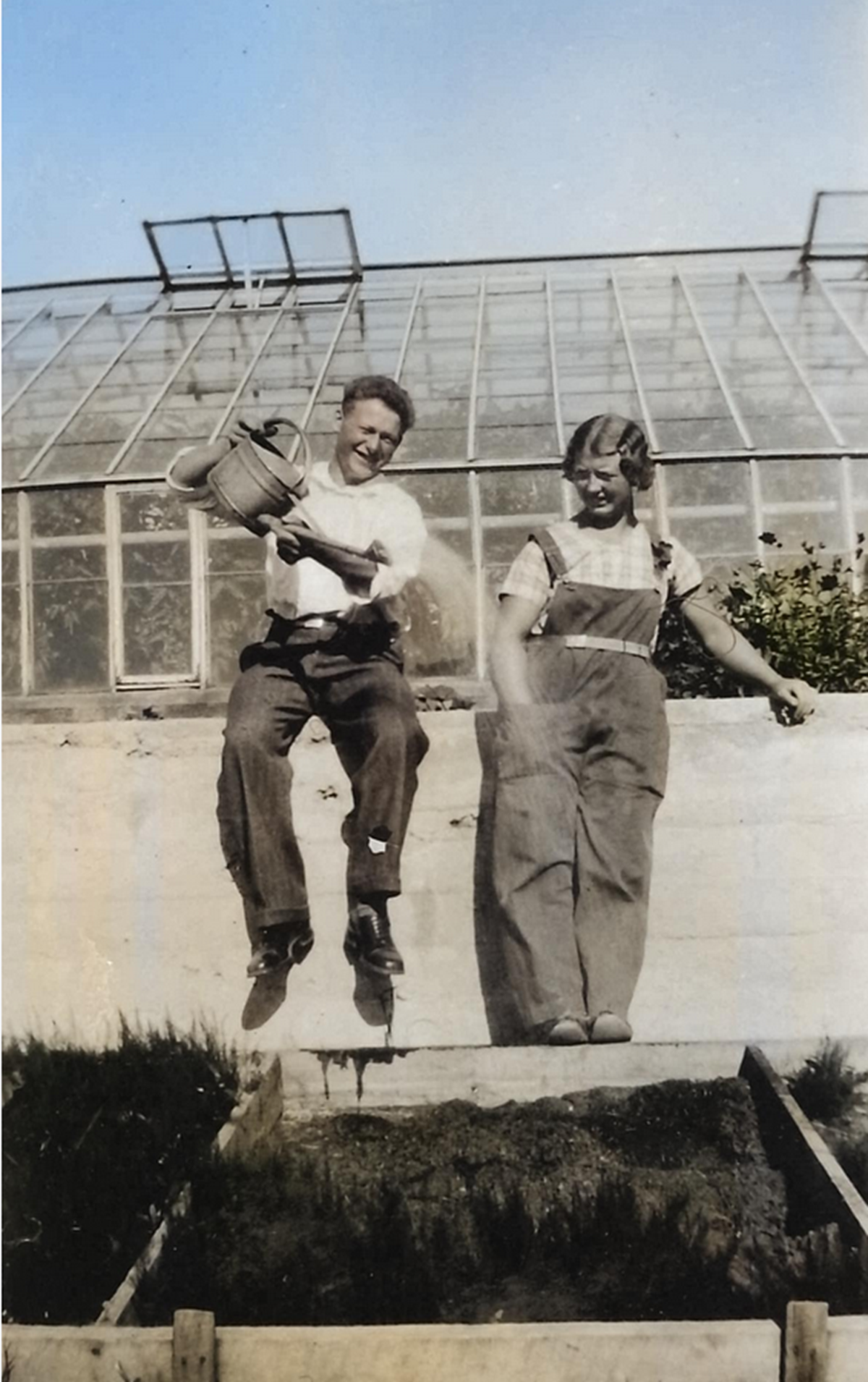 Bilde av to glade gartnerskoleelver. De sitter på kanten foran drivhusene og heller vann fra en kanne i en plantekasse.
Bildet er fra 1933.