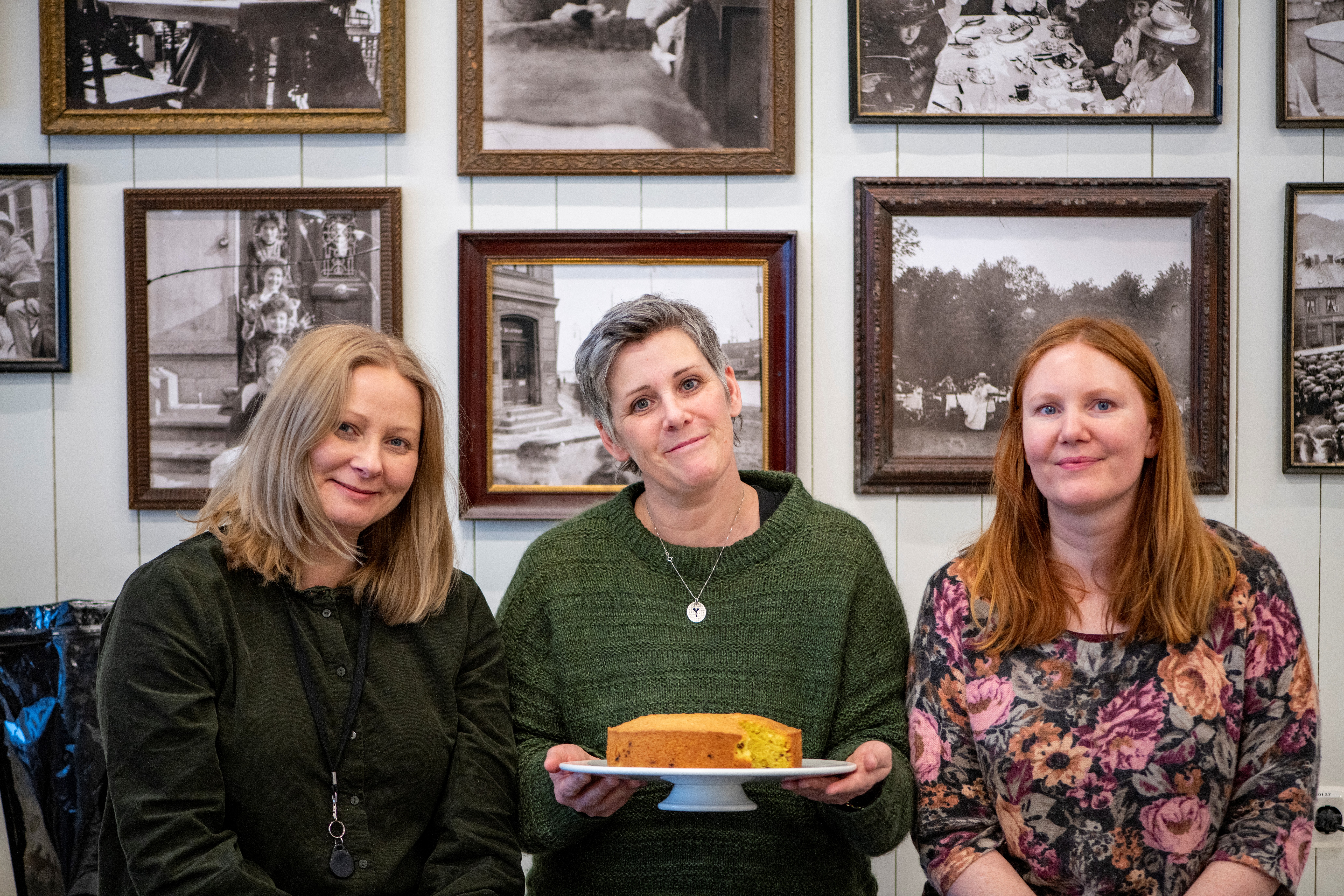 Tre kvinner sitter sammen. Kvinnen i midten holder frem et kakefat med en kake på.